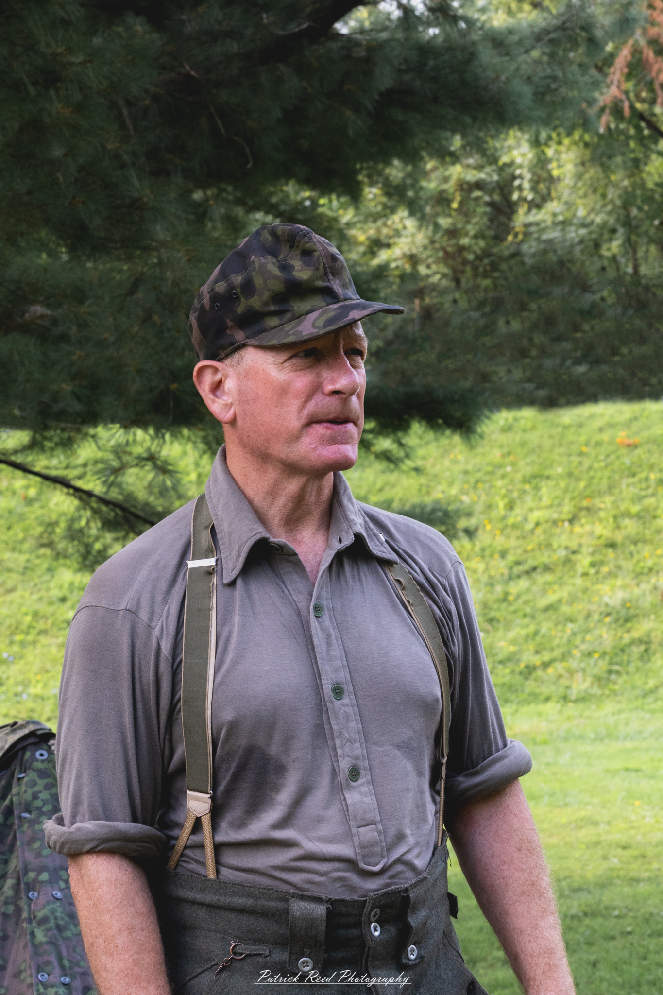 A German soldier wearing a camouflage cap, standing alert and focused. The soldier's uniform and gear blend into the environment, emphasizing the tactical nature of his role in the field.